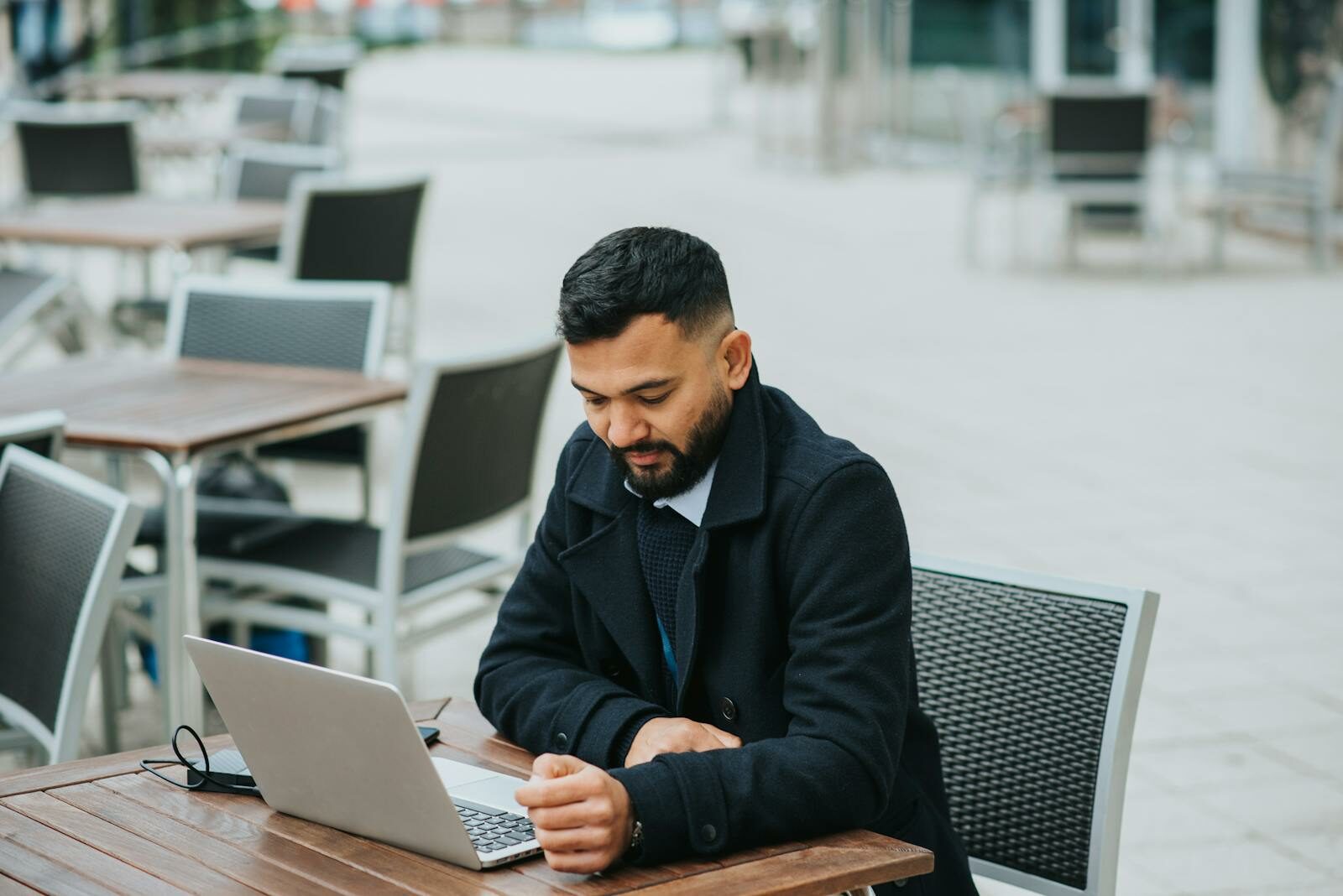 Young man working on a laptop at an outdoor café table, appearing focused and thoughtful.